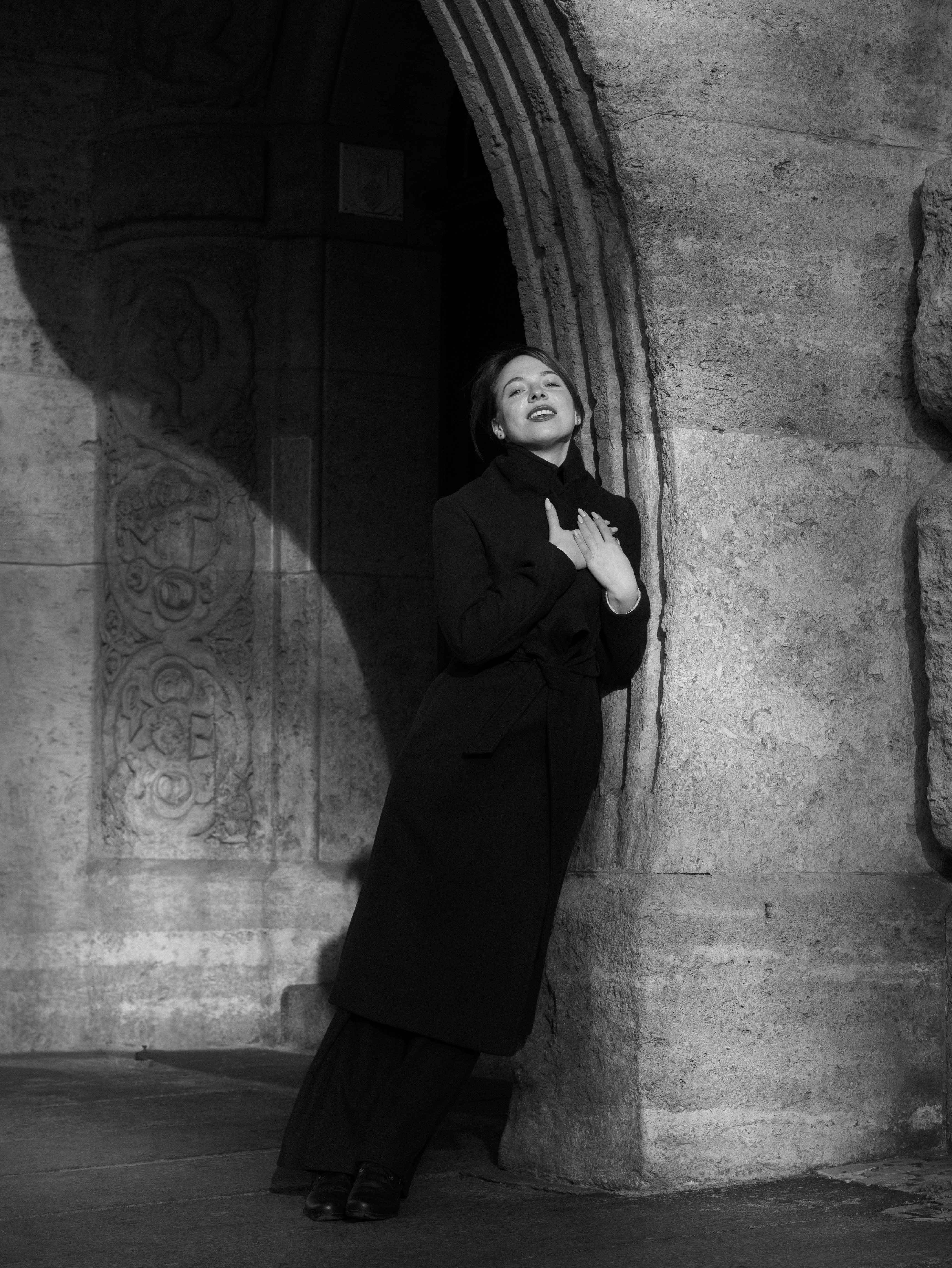 Black-and-white street-style photo of a woman leaning on a wall with shadows echoing her pose, wearing a long black coat and lifting her chin while holding the collar