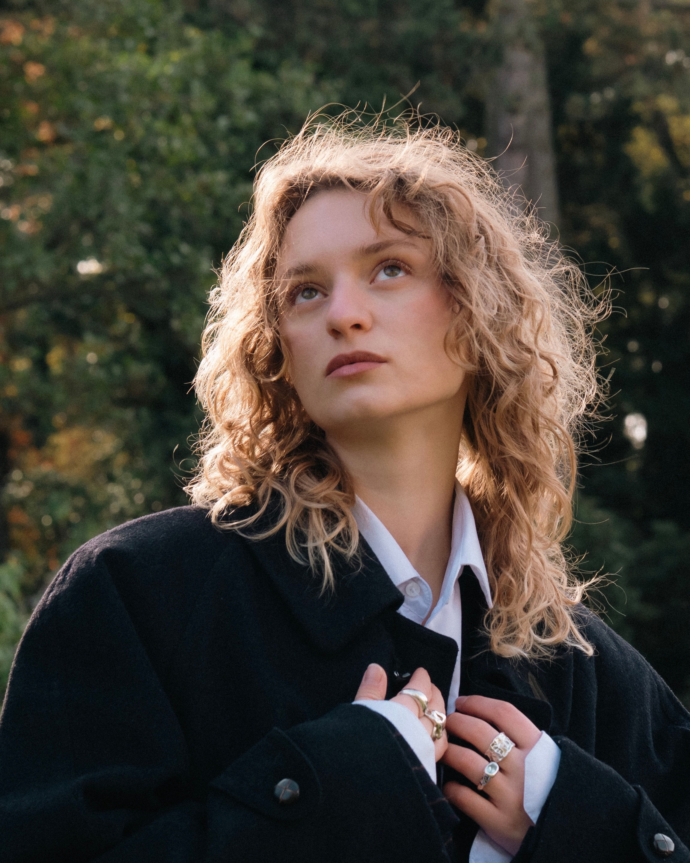 Outdoor portrait of a blonde woman with curls looking upward against a forest background, backlit by the sun and wearing a black coat
