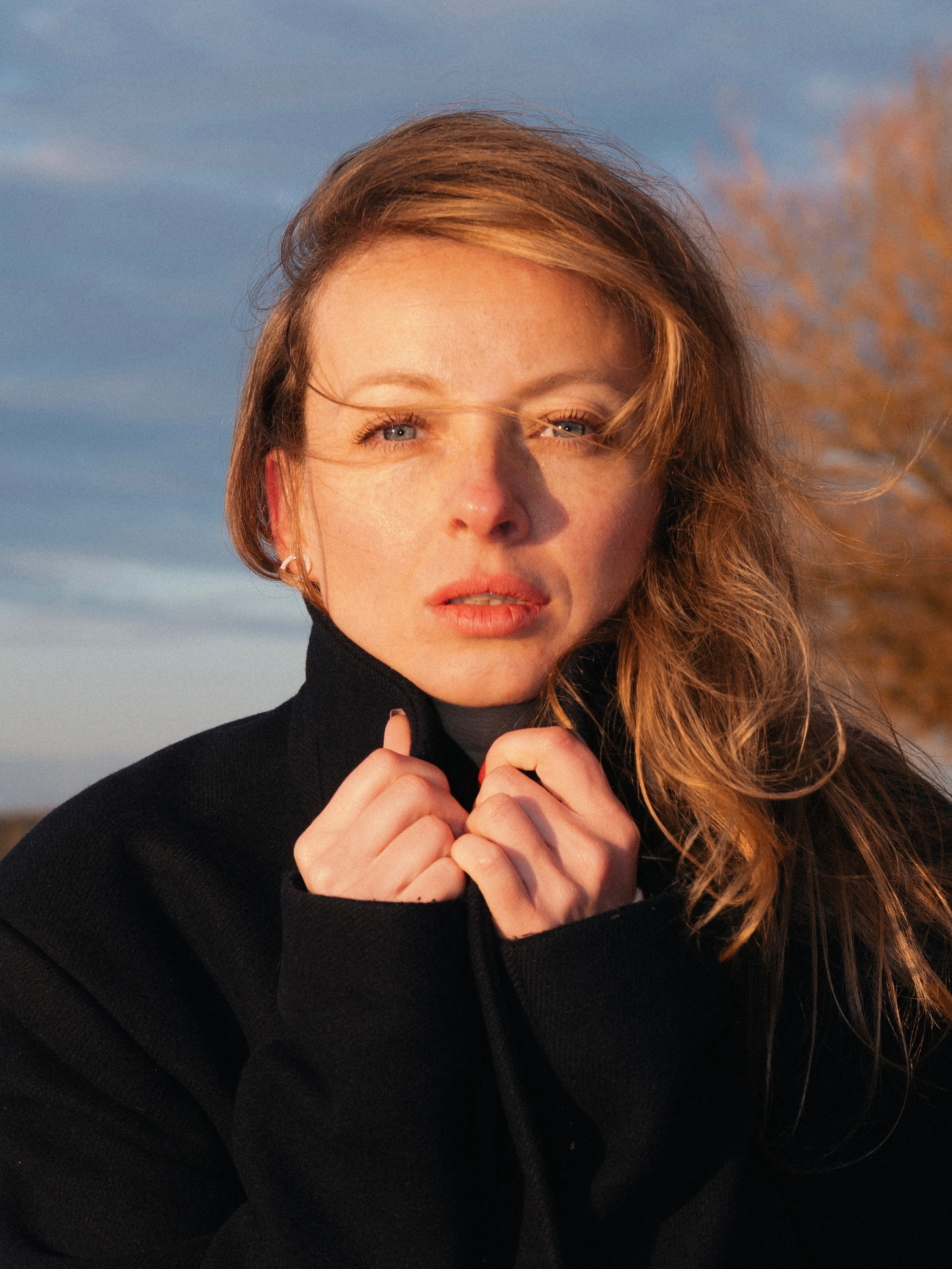Golden-hour color portrait of a blonde woman with blue eyes in a black coat, holding the collar with both hands