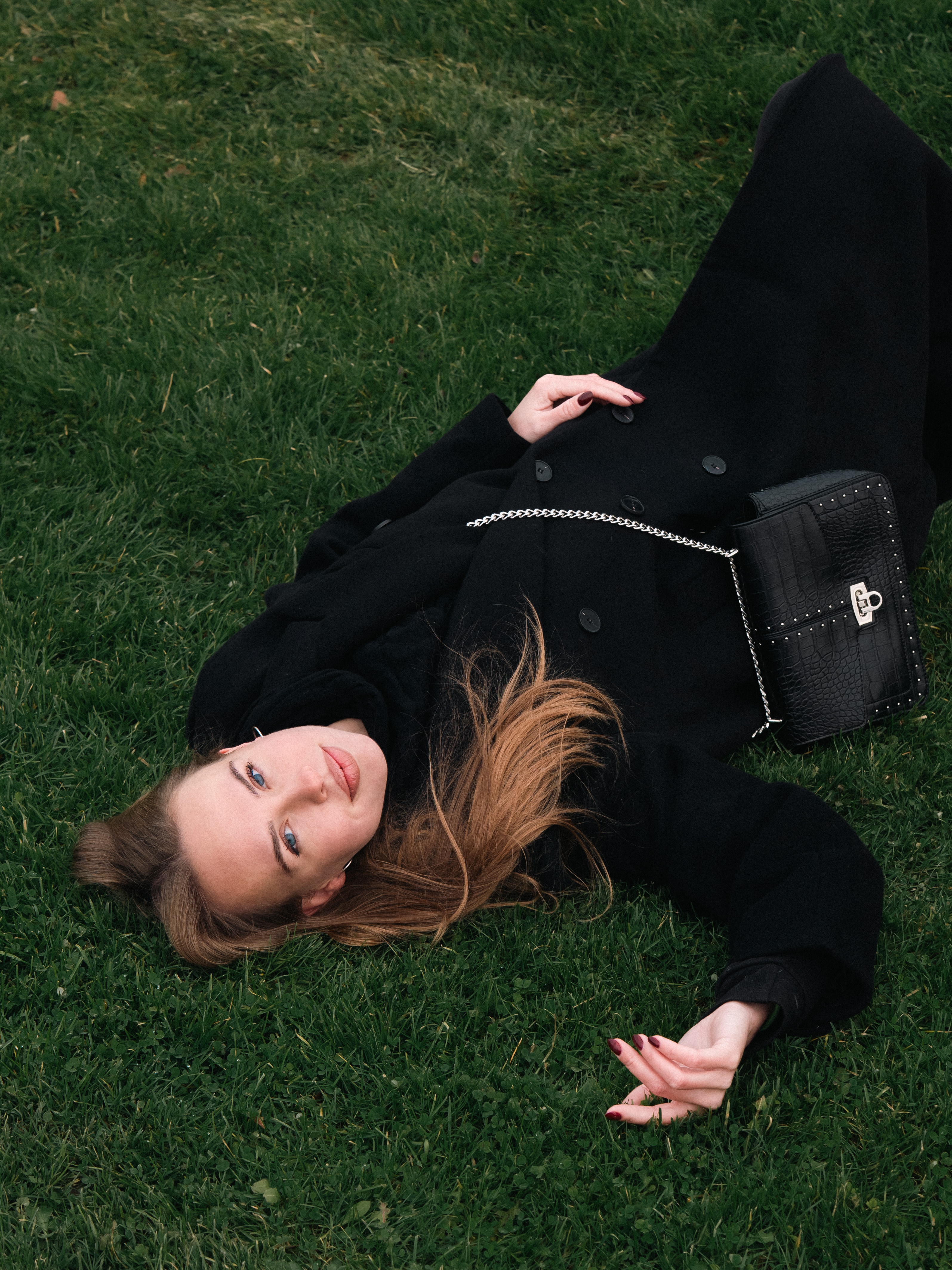 Blonde woman lying upside down on green grass wearing a black coat and a black cross-body purse with a silver chain