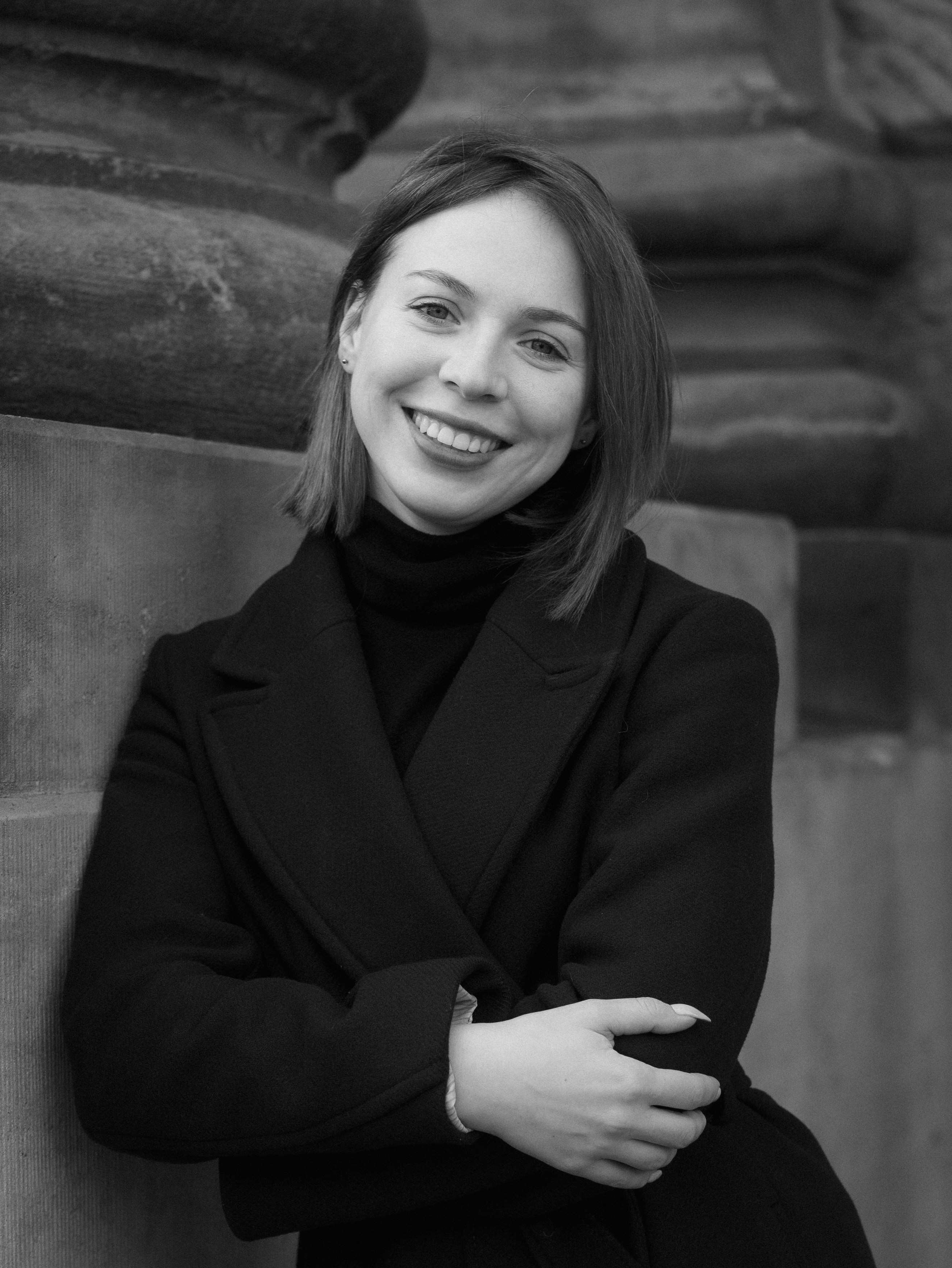 Black-and-white street-style portrait of a smiling woman leaning on a wall with her arms crossed, wearing a black coat