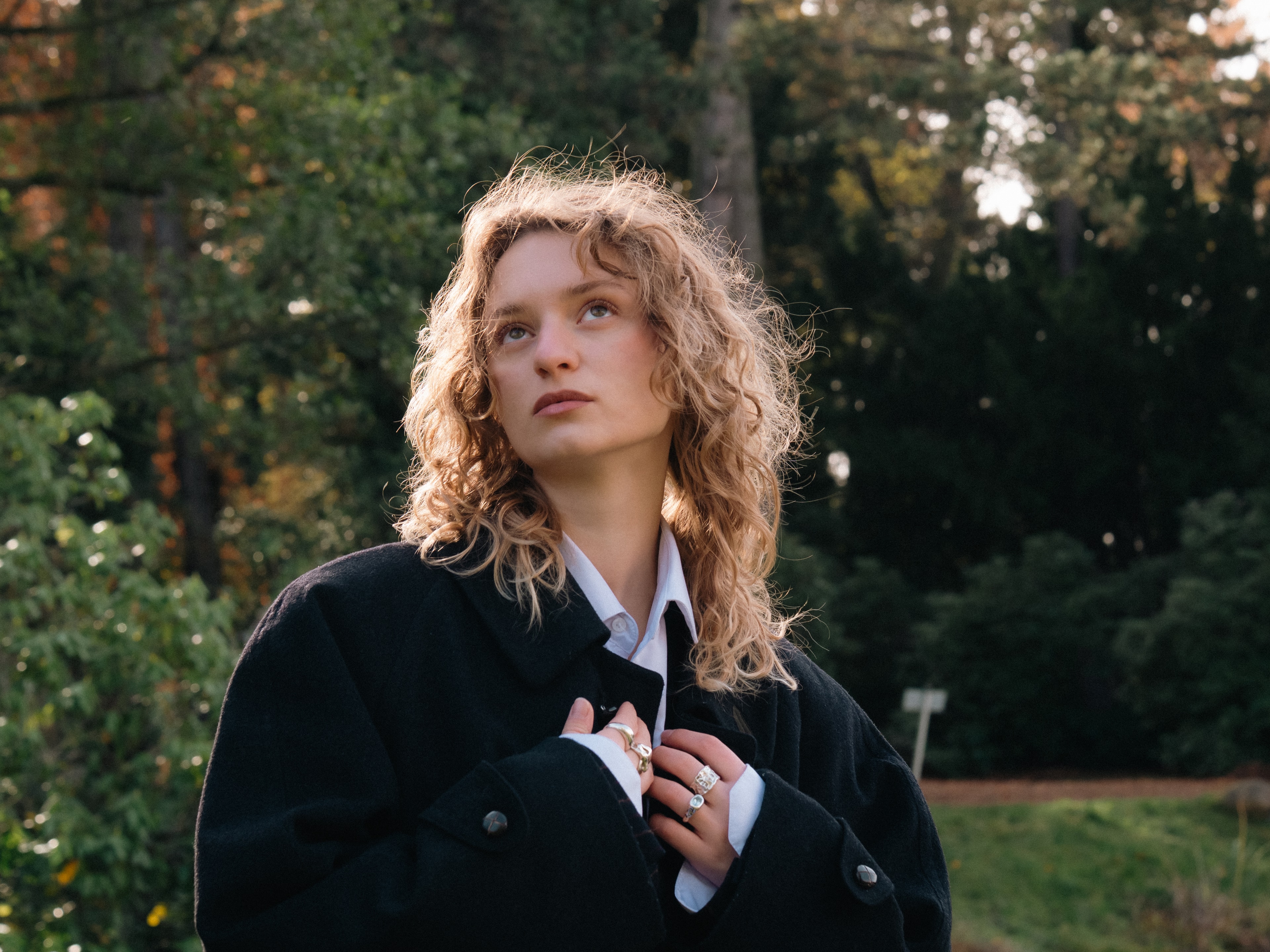 Outdoor portrait of a curly-haired woman in a dark coat looking upward in forest light