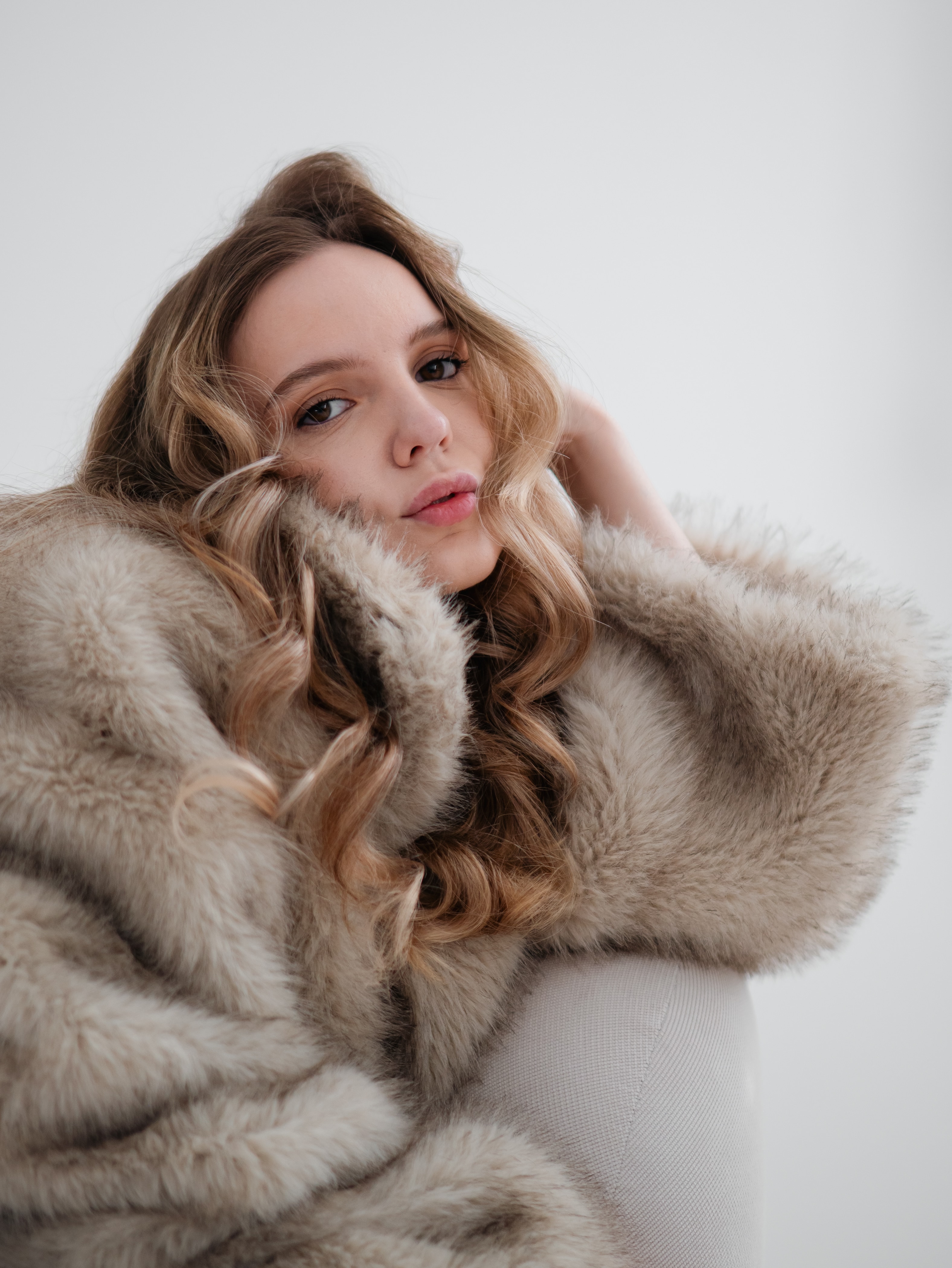 Soft portrait of a blonde woman wrapped in a light fur coat against a pale background