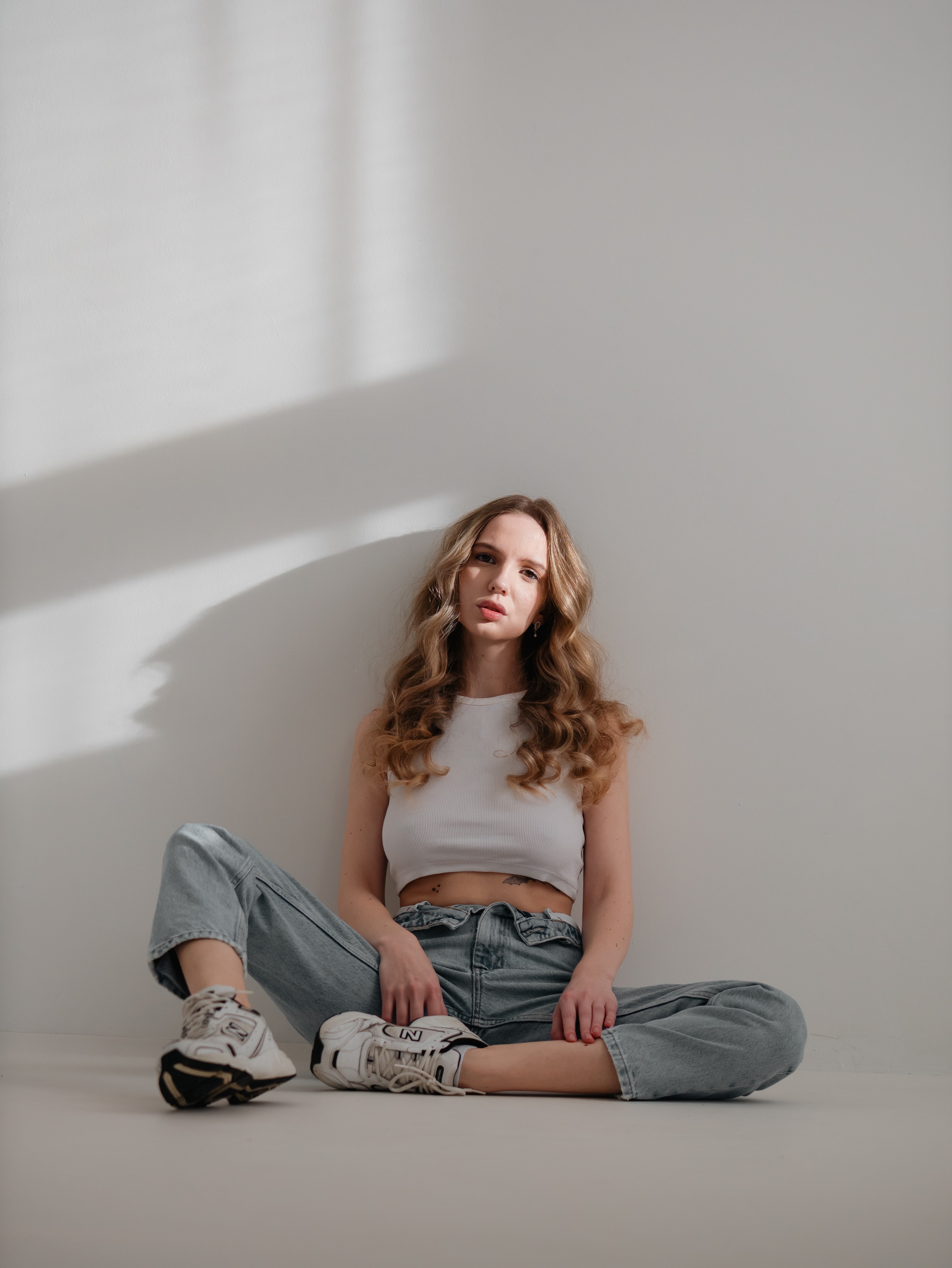 Full-body studio portrait of a woman seated on the floor against a white wall with a long shadow