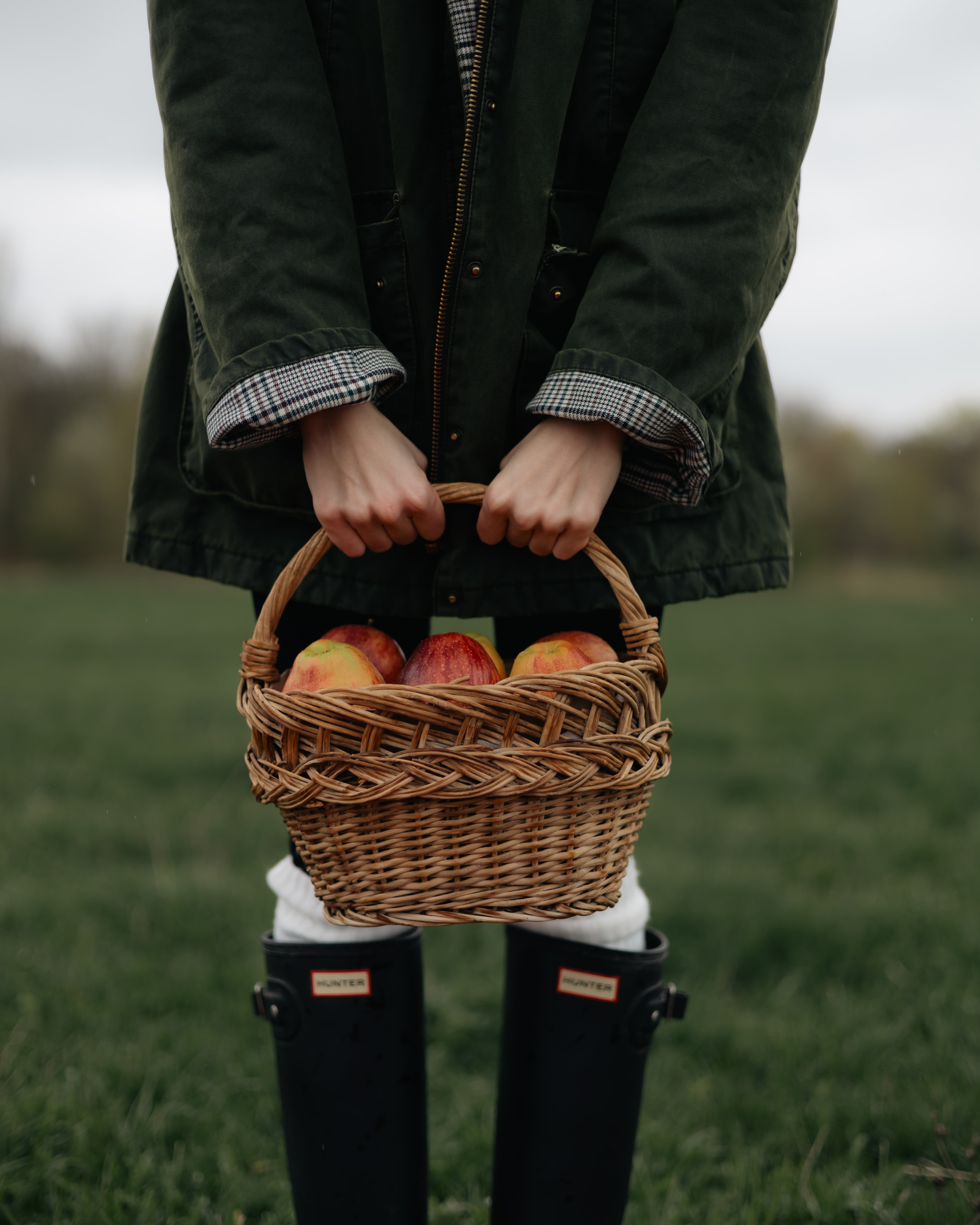 Person holding a wicker basket of apples in a field while wearing rain boots